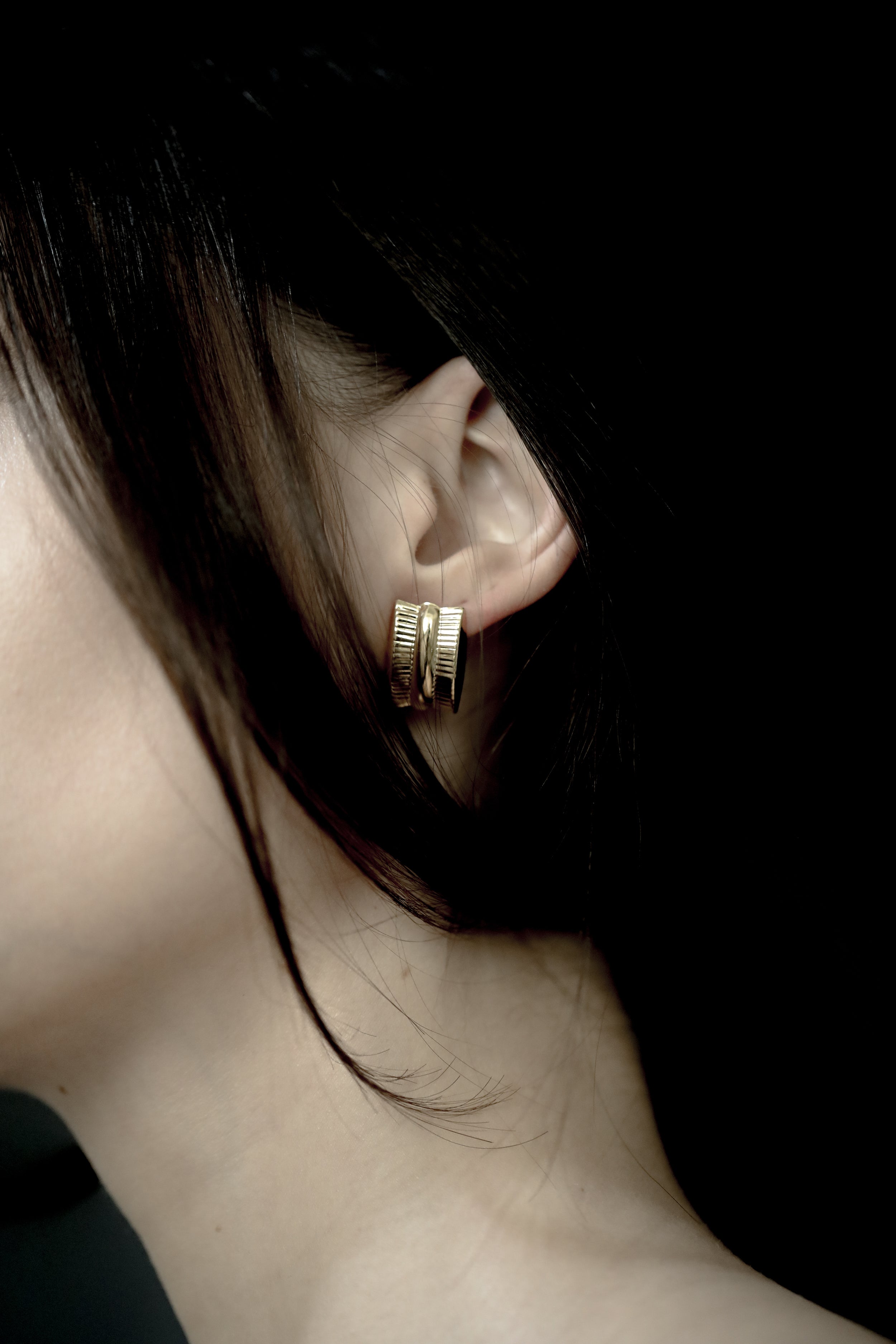 Close-up of a brunette wearing the AB Ellie gold ribbed channel earring with a dark background