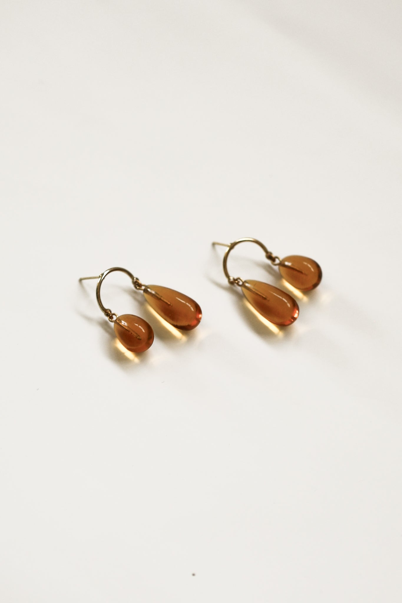 Flat image of glass asymmetric drop earrings in gold and amber glass by Agytha on white background