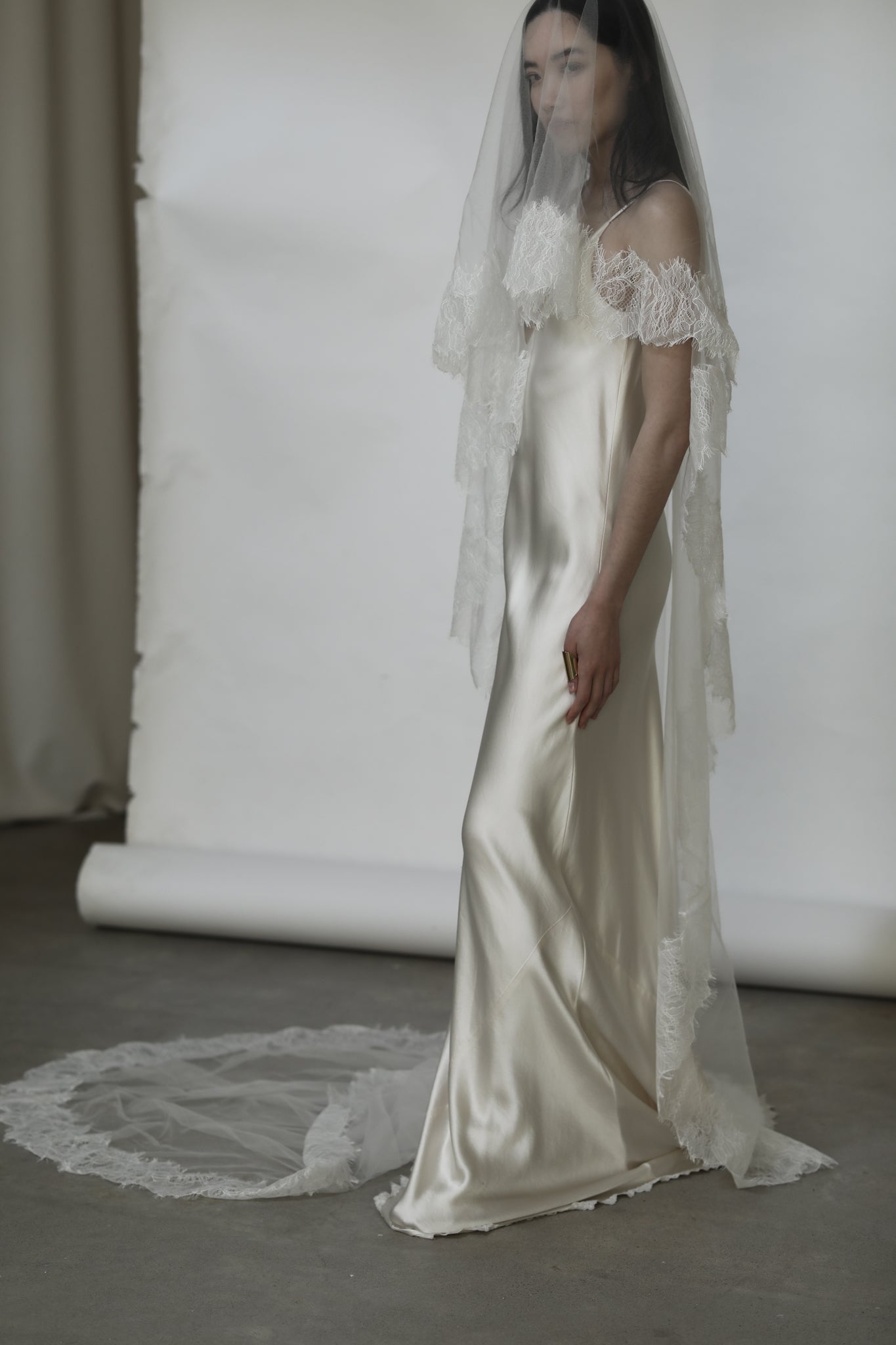 Dark haired bride wearing a silk wedding dress with a long train and lace veil against a white background