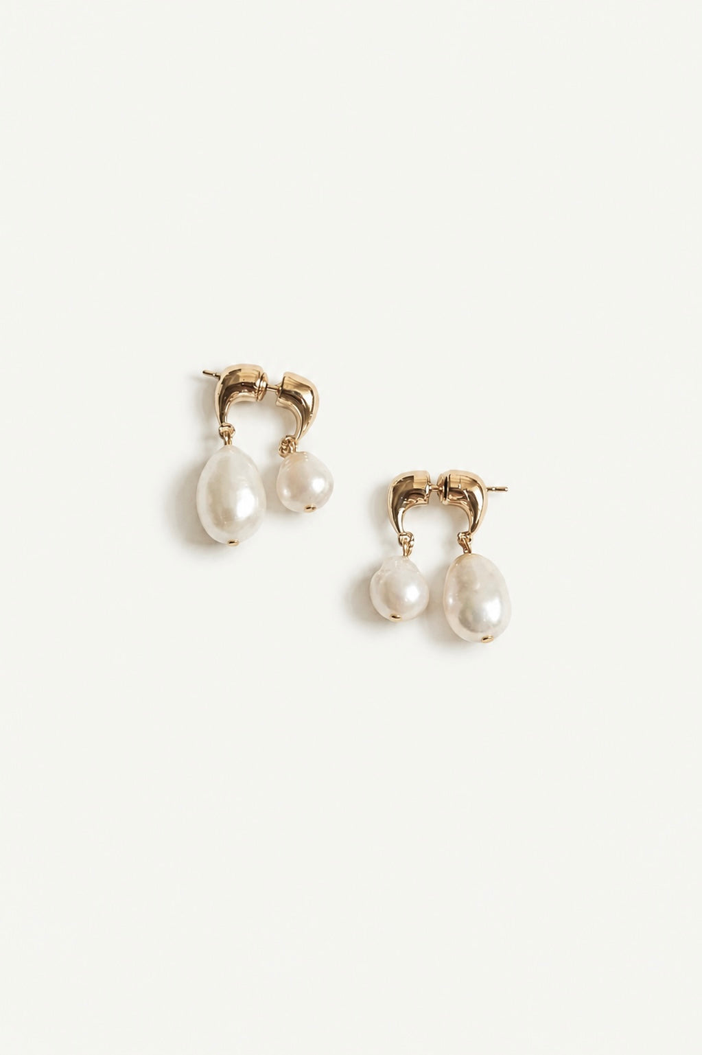 A pair of gold-tone drop earrings featuring irregular white baroque pearls, displayed on a clean, off-white background.