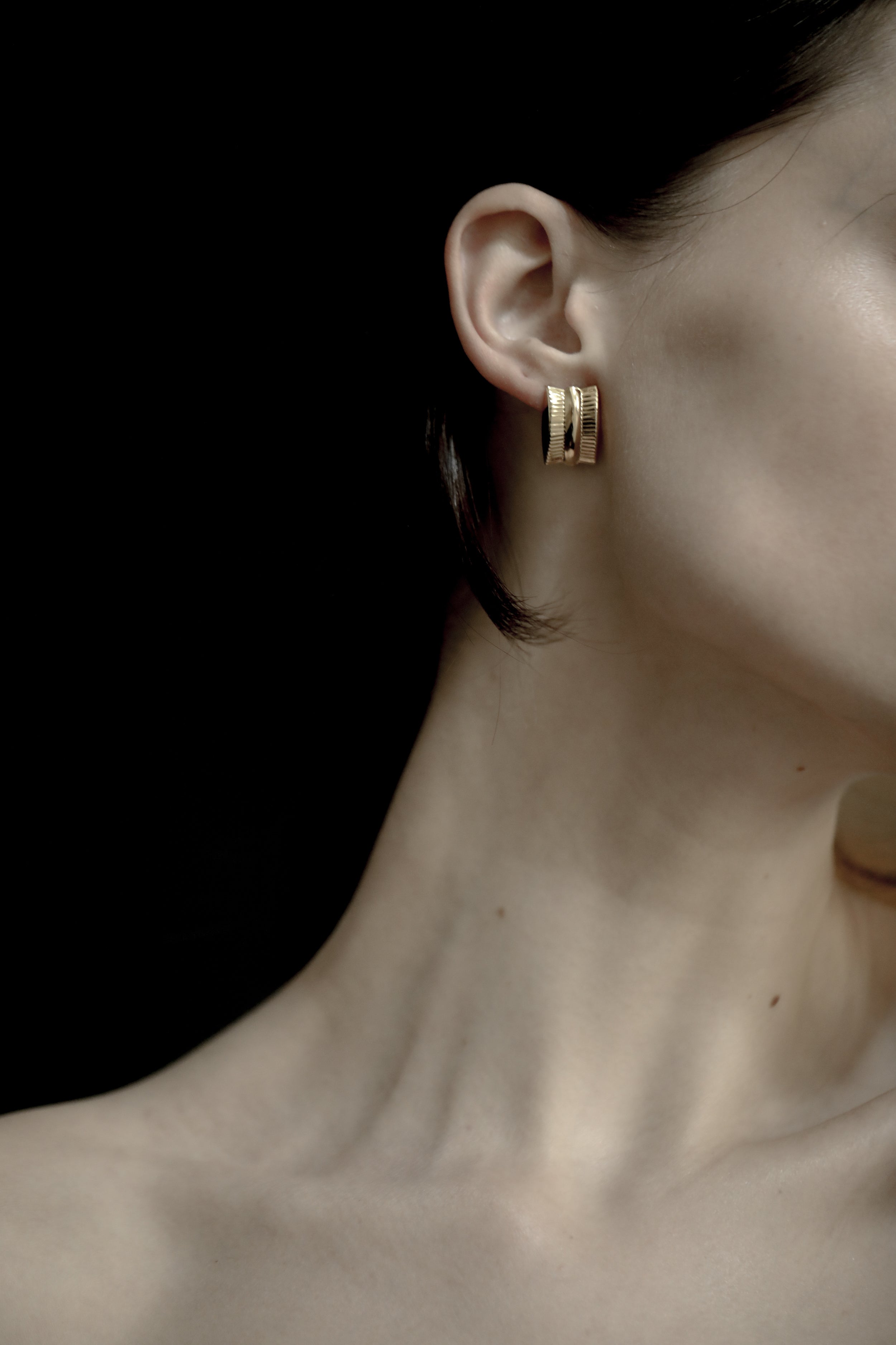 Close-up of an brunette woman wearing a the AB Ellie ribbed gold hoop earring against a dark background