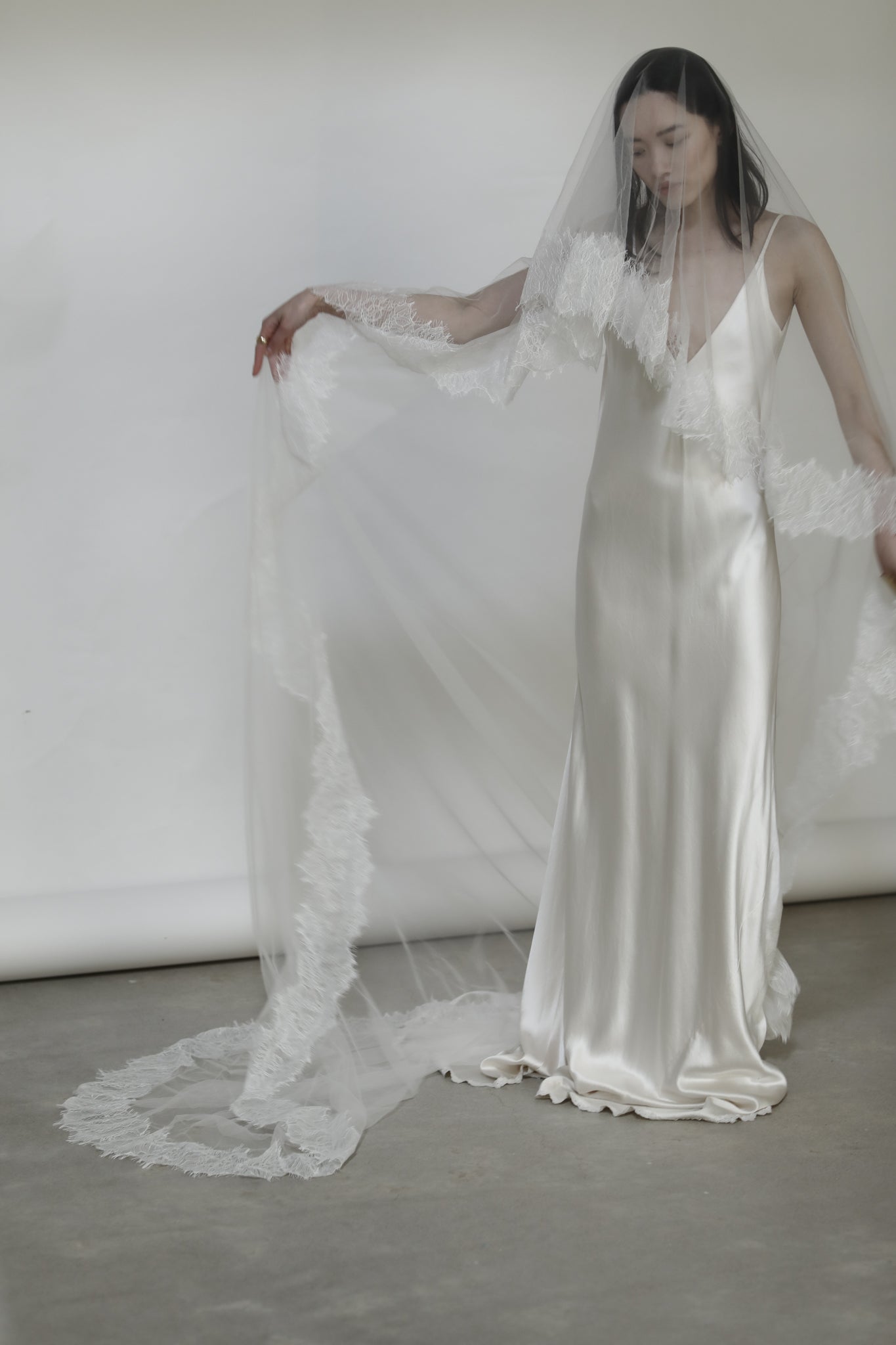 Woman in a ivory silk wedding dress holding a long white veil with lace details against a white background.